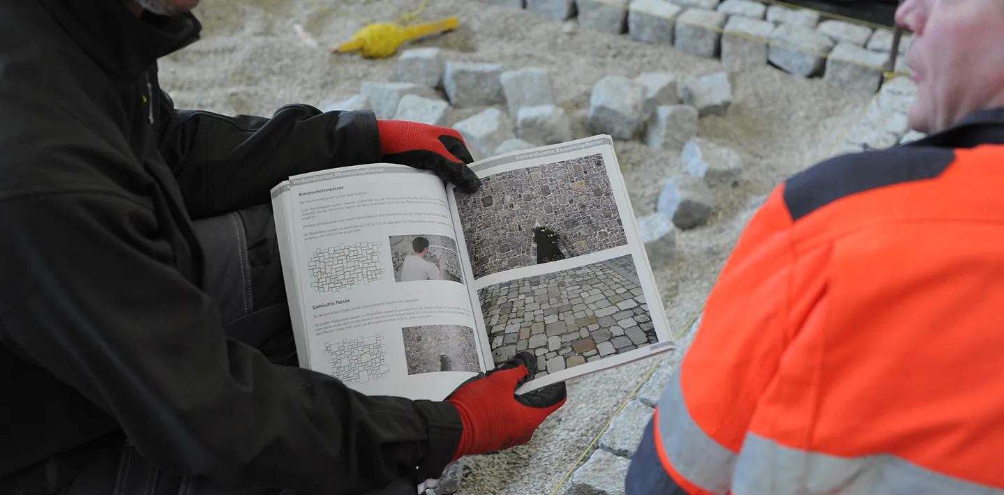 Zwei Bauarbeiter in Arbeitskleidung blättern an einer Pflasterbaustelle in einem aufgeschlagenen Fachbuch mit Verlegemustern für Kleinsteinpflaster