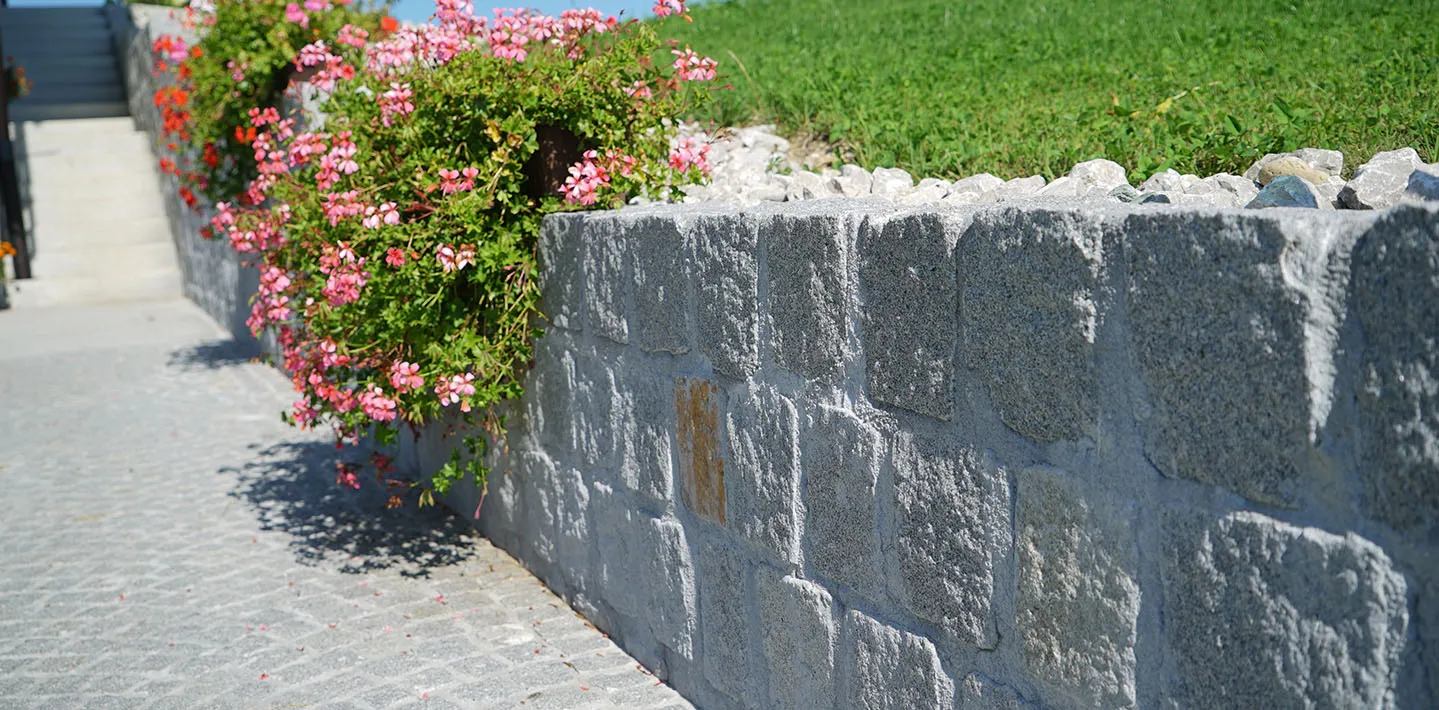 Mauer aus grob bearbeiteten Granitsteinen entlang eines gepflasterten Weges, darüber blühende Geranien und Rasen