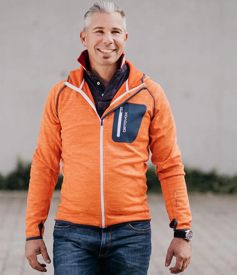 Mario Tomasek in oranger Fleecejacke steht auf einer gepflasterten Fläche vor einer Betonwand und lächelt in die Kamera