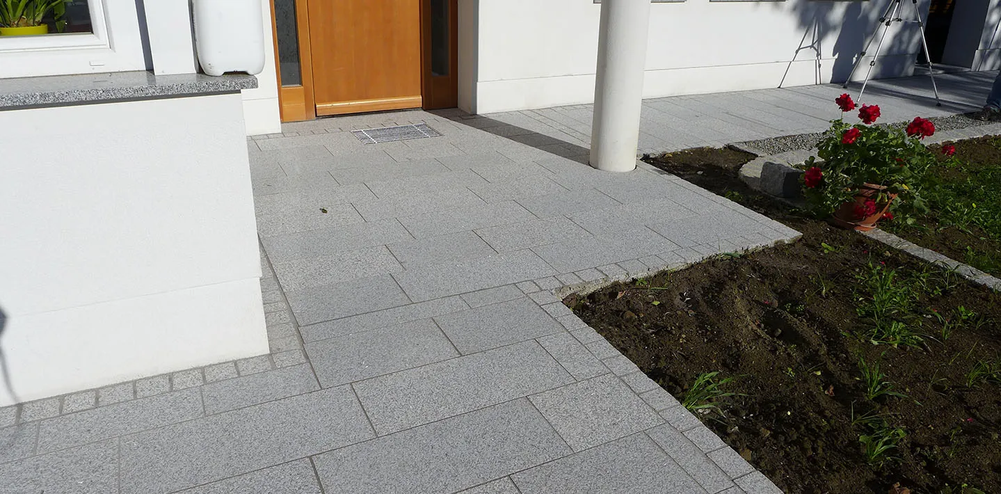 Frisch verlegter Granit-Plattenbelag vor einem Hauseingang mit Entwässerungsrinne vor der Tür, daneben ein noch offenes Beet