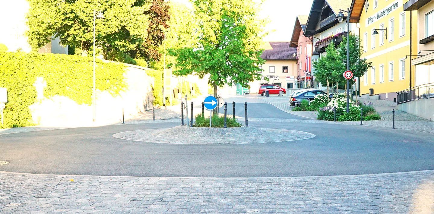 Ortszentrum mit gepflastertem Kreisverkehr aus Granit-Kleinstein zwischen Asphaltfahrbahnen, im Morgenlicht, mit Häuserzeile und Baumreihe im Hintergrund