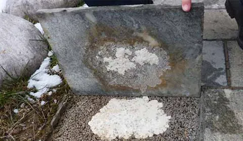 Angehobene Granitplatte, an deren Unterseite eine weiße kristalline Ausblühung haftet; dieselbe Ablagerung liegt darunter im Splittbett