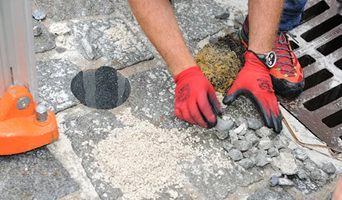 Neben einem Straßeneinlauf ist die Pflasterfläche geöffnet: Hände in roten Arbeitshandschuhen nehmen die Steine ab und legen Bettung und Fugenmaterial frei