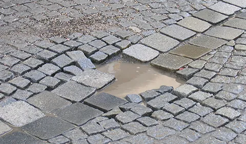 Eingesunkene Stelle in einer Granit-Kleinsteinfläche, in der sich trübes Regenwasser sammelt; die umliegenden Steine stehen schief und lose