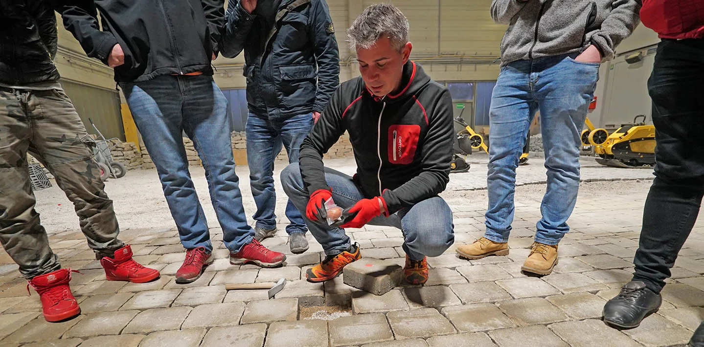 Mario Tomasek kniet in einer Schulungshalle vor einer Pflasterfläche und erklärt einer Gruppe von Teilnehmern den Steinversatz