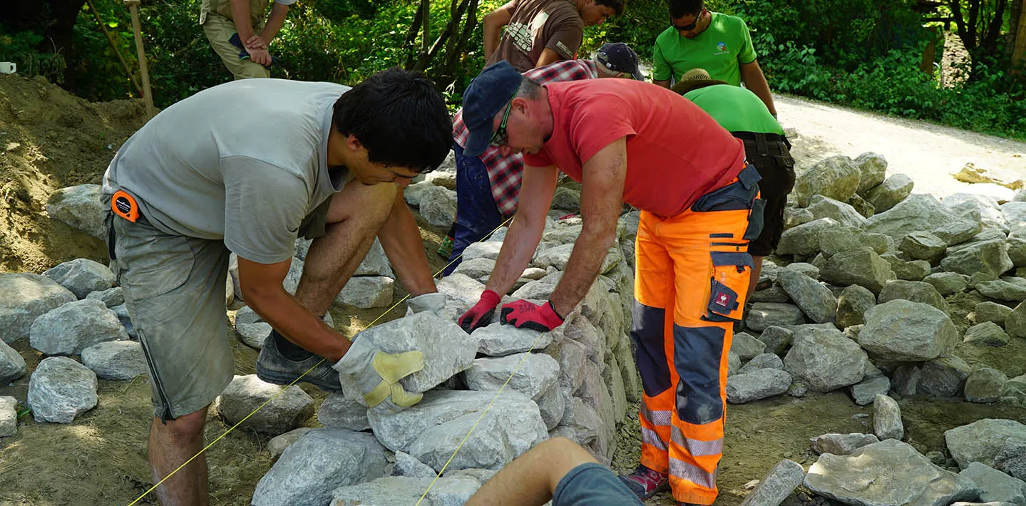 Eine Gruppe von Seminarteilnehmern setzt im Freien gemeinsam eine Trockensteinmauer, ein Teilnehmer legt einen Bruchsteinquader entlang einer gespannten Schnur ein